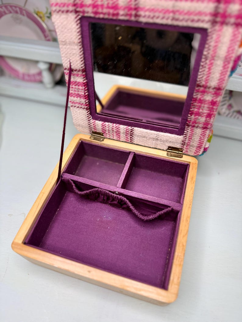 May include: Open wooden jewelry box with a purple interior and a plaid fabric lid. The lid has a built-in mirror. The box is open, revealing compartments for storing jewelry. The fabric on the lid is pink, white, and purple.