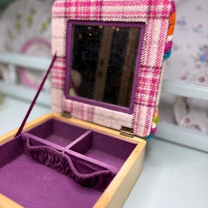 May include: Open wooden jewelry box with a purple interior and a plaid fabric lid. The lid features a mirror and is trimmed with colorful pom-poms. The box is open, revealing compartments for storing jewelry.