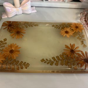 May include: Rectangular resin tray with embedded orange daisies and fern-like leaves. The tray has a light yellow background and a clear, glossy finish. The edges of the tray are a warm brown color. A decorative item for the home.