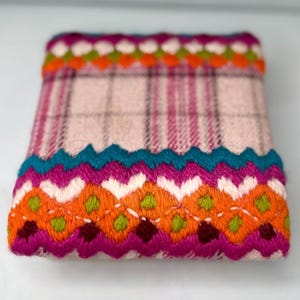 May include: A small, rectangular textile item with a pink plaid pattern. The edges are adorned with vibrant, embroidered trim in shades of orange, teal, magenta, and green. The trim features a zig-zag design.