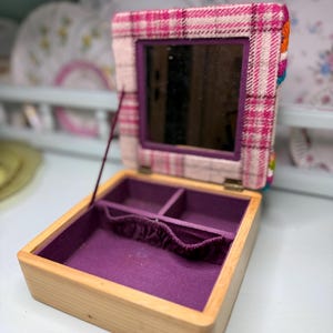 May include: Open wooden jewelry box with a purple velvet interior and a plaid fabric lid. The lid features a small mirror and is trimmed with pink, white, and orange fabric. The box is open, revealing compartments for storing jewelry.