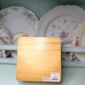 May include: A square, light-colored wooden coaster with a visible wood grain. A white label with black text and a barcode is affixed to the coaster. The background features a collection of decorative plates.