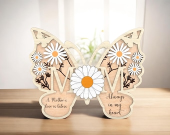 Op de afbeelding: Houten vlinderuitsparing met het woord "MOM" in het midden. De vlinder heeft witte madeliefjes en bruine stelen. De tekst "A Mother's love is timeless" staat aan de linkerkant en "Always in my heart" aan de rechterkant.