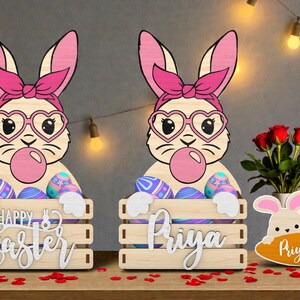 May include: Wooden Easter bunny decorations. Two bunnies with pink bows, heart-shaped glasses, and bubblegum bubbles. Each holds a crate with Easter eggs. One says "Happy Easter", the other "Priya". A small bunny with a carrot is also present.