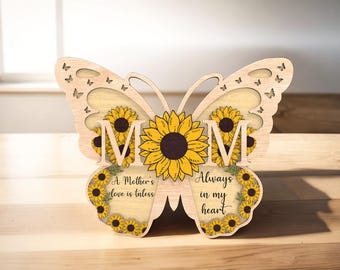 Op de afbeelding: Houten vlinderuitsparing met een zonnebloemontwerp en het woord "MOM" in het midden. De vlinder heeft de tekst "A Mother's love is timeless" en "Always in my heart" op de vleugels.