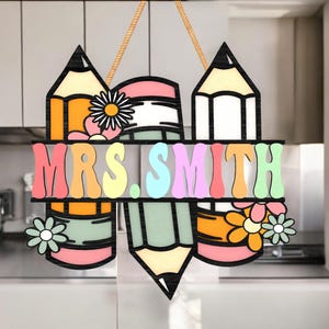 Puede incluir: Un letrero de madera con el nombre "MRS. SMITH" en letras retro, rodeado de diseños de lápices y flores. El letrero está suspendido por una cuerda. La combinación de colores incluye naranja, rosa, amarillo y verde, creando una pieza vibrante.