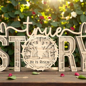 May include: Wooden sign with the words "true STORY" in a layered design. The letter "O" features a circular design with a figure and the text "He Is Risen". The sign is set on a wooden surface with a blurred background of greenery and flowers.