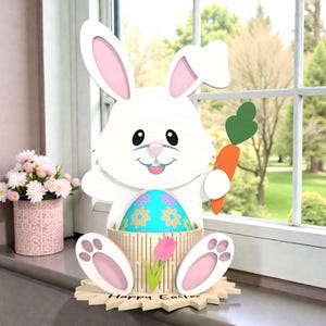 Può includere: Un coniglietto pasquale di legno bianco che tiene in mano una carota e un uovo con un disegno floreale. Il coniglietto è davanti a una finestra, con le parole "Happy Easter" alla base. A sinistra c'è un vaso di fiori rosa con fiori rosa.