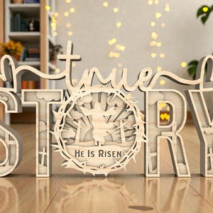 May include: A wooden sign that spells out "true STORY" in a layered design. The letter "O" features a circular design with a figure and the text "He Is Risen". The sign is light brown and sits on a wooden surface.