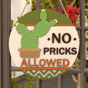 May include: A round wooden sign with a cactus illustration and the words "NO PRICKS ALLOWED." The sign features a green cactus in a brown pot, set against a light background with a green and orange border. A rope is attached for hanging.