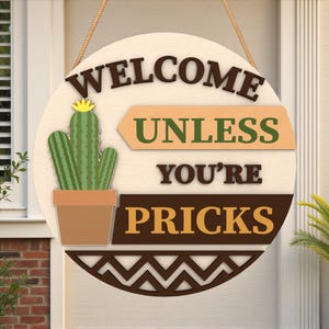 May include: Round wooden welcome sign with a cactus illustration and the text "WELCOME UNLESS YOU'RE PRICKS." The sign features a light brown background, a brown cactus pot, and a decorative zig-zag border.