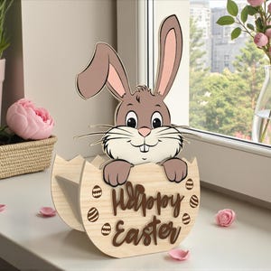 May include: A wooden Easter decoration featuring a cartoon bunny peeking out of a cracked egg. The egg has the words "Happy Easter" and Easter egg designs. The bunny is brown and tan with pink inner ears. The decoration is on a white surface.