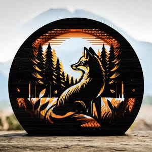 Könnte beinhalten: Runde Holz-Kunst mit einer Fuchssilhouette in einer Waldszene. Der Fuchs ist in Orange- und Gelbtönen vor schwarzem Hintergrund dargestellt, mit einer Bergkette im Hintergrund. Das Kunstwerk befindet sich auf einer Holzoberfläche.