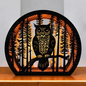May include: A black wooden owl silhouette lamp with a forest scene. The owl is perched on a branch, surrounded by trees, with an orange glow emanating from within. The lamp is on a wooden surface.