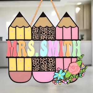 Op de afbeelding: Een houten lerarenbord met drie potloodvormen en de naam "MRS. SMITH" in kleurrijke letters. De potloden zijn geel, roze en hebben een luipaardprint. Een bloemstuk met een roze roos en blauwe bloemen bevindt zich aan de rechterkant.