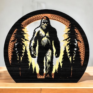 May include: A black wooden Bigfoot silhouette art piece. The design features a detailed depiction of Bigfoot in a forest setting, with trees and a circular frame. The artwork is set against a warm, golden background.