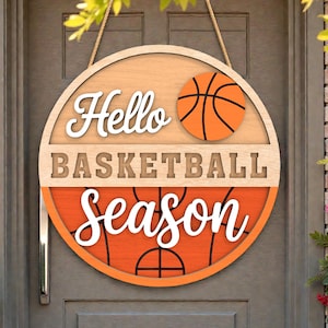 May include: Round wooden sign with the words "Hello Basketball Season" in white script. The sign features a basketball graphic and is designed with orange, tan, and brown colors. It hangs from a tan rope.