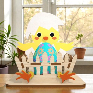 Może przedstawiać: Drewniana dekoracja wielkanocna przedstawiająca żółte pisklę wychodzące z białej skorupki jajka, z niebieskim jajkiem ozdobionym kwiatowymi wzorami. Pisklę umieszczone jest w drewnianym płocie, z pomarańczowymi stopami u podstawy.