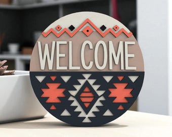 Puede incluir: Un letrero redondo de madera con la palabra "WELCOME" en blanco. El fondo es beige, con un diseño geométrico en coral y negro en la parte superior y un patrón azul marino y coral en la parte inferior. Decoración del hogar.