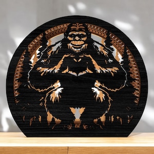 May include: A circular wooden artwork featuring a Bigfoot design. The piece has a layered construction with a black exterior, a brown middle section, and a detailed Bigfoot figure at the centre. It is displayed on a wooden surface.
