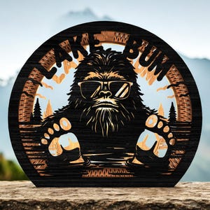 May include: A circular wooden sign with a Bigfoot design wearing sunglasses, and the text "LAKE BUM". The sign has a layered design with a black outer frame and a light brown inner layer, depicting a lake scene. The sign is made of wood.