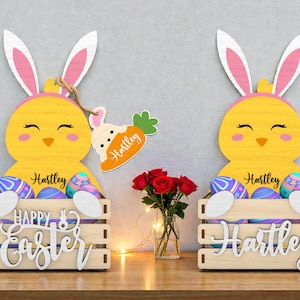 May include: Two wooden Easter decorations shaped like yellow chicks with bunny ears, holding a crate of Easter eggs. The crates have the words "Happy Easter" and a name. A carrot-shaped tag hangs from one. A vase of red roses sits nearby.