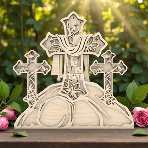 May include: A wooden Easter decoration featuring three crosses and the text "He is Risen." The crosses are intricately cut with floral designs. The decoration is set against a blurred background of greenery and pink roses.