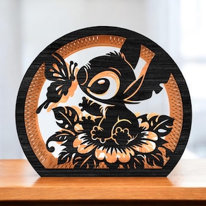 Peut inclure: Une pièce décorative en bois superposée représentant une silhouette noire d'un personnage de dessin animé aux grands yeux, assis sur une fleur, avec un papillon. La pièce est encadrée par une bordure circulaire noire et marron.