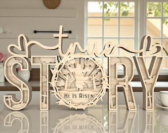 May include: Wooden sign with the words "true STORY" in a layered design. The letter "O" features a circular cutout with a religious scene and the text "He Is Risen". The sign is made of light-colored wood and is displayed on a reflective surface.