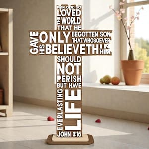 May include: A wooden cross with the text of John 3:16. The cross is brown with white lettering and stands on a rectangular base. The cross is in a room with a window and a bookshelf.
