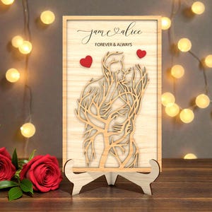 Könnte beinhalten: Helle Holzplakette mit einem lasergeschnittenen Design eines sich küssenden Paares, mit den Namen "jam & alice" und dem Satz "Forever & Always". Zwei kleine rote Herzen sind enthalten. Die Plakette wird auf einem Holzständer präsentiert.