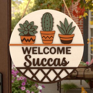 Puede incluir: Cartel de madera redondo con fondo crema, con tres suculentas en macetas sobre las palabras "WELCOME Succas" en marrón. El cartel tiene un diseño de celosía marrón en la parte inferior y está suspendido por una cuerda natural.