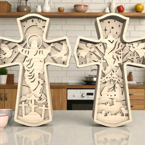 May include: Two wooden crosses with intricate layered designs. One cross features a figure with outstretched arms and doves, the other depicts a landscape with trees and figures. Both are light-colored and stand on a countertop.