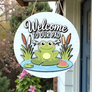 May include: Round wooden sign with the text "Welcome to our Pad". The sign features a cartoon frog on a lily pad, cattails, dragonflies, and a pink water lily. The background is light blue.