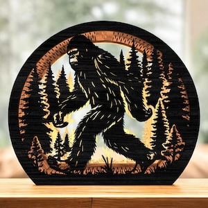 May include: A black wooden Bigfoot silhouette walking through a forest scene, framed in a circular design. The artwork features detailed cutouts of trees and the creature, creating a layered effect. The piece is displayed on a wooden surface.