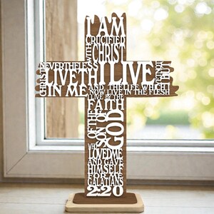 May include: A wooden cross with the text "I am crucified with Christ" and other religious text cut out. The cross is brown with white lettering and stands on a light brown base. The background is a window.