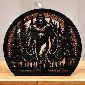 May include: A black wooden Bigfoot silhouette art piece. The design features a detailed cutout of Bigfoot in a forest scene, with trees and a circular frame. The piece is set on a wooden surface.
