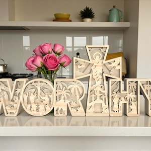 Könnte beinhalten: Dekorative Holzbuchstaben, die das Wort "WORTHY" bilden, mit aufwendigen Ausschnitten, die religiöse Szenen darstellen. Die hellen Buchstaben stehen auf einer Arbeitsplatte, mit einer Vase mit rosa Rosen im Hintergrund.