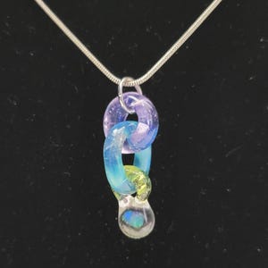 May include: A silver chain necklace with a pendant featuring three linked, colorful glass rings. The top ring is purple, the middle is blue, and the bottom is green with a clear, teardrop-shaped glass bead.