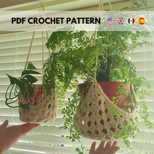 Pode incluir: Dois suportes de plantas de crochê suspensos com plantas. Os suportes de crochê são bege e suportam vasos de terracota cheios de plantas verdes. A imagem inclui o texto "PDF CROCHET PATTERN" e bandeiras dos Estados Unidos, Reino Unido, Itália e Espanha.