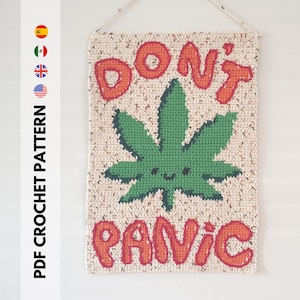 Op de afbeelding: Een wandkleed van gehaakt garen met de woorden "Don't Panic" in oranje en een groen marihuanablad met een glimlachend gezicht in het midden.