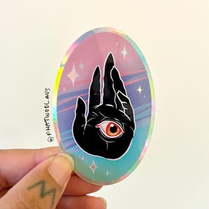May include: A holographic sticker with a black hand with an eye in the palm. The background is a pink, blue, and purple gradient with stars.