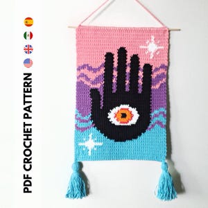 May include: A crochet wall hanging with a black hand with an orange eye in the center. The hand is surrounded by pink, purple, and blue waves. The hanging has two blue tassels.