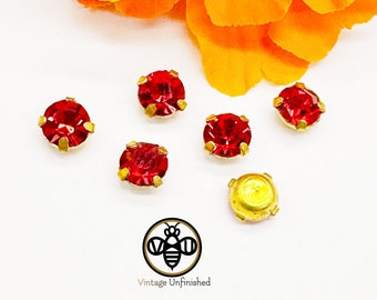 May include: Six small round red rhinestones set in gold-toned metal prongs, arranged near a bright orange marigold flower. One rhinestone is flipped over to show its smooth gold-toned metal back. A logo with a bee and the text Vintage Unfinished.