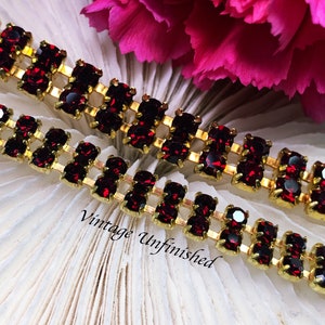 May include: A gold-toned metal chain featuring two parallel rows of deep red faceted stones set in prongs. The text "Vintage Unfinished" is printed in black script across the center of the image, positioned over the jewelry and white texture.