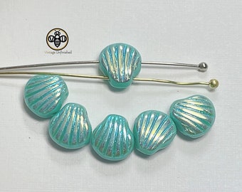 May include: Seven iridescent turquoise glass beads shaped like seashells. The beads are arranged in a semi-circle on a white surface.