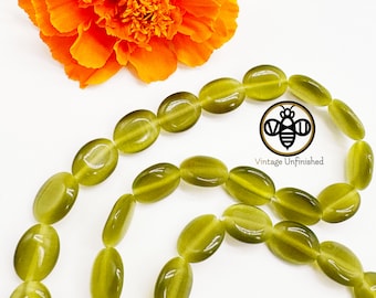 May include: A strand of oval-shaped green gemstone beads. The beads are smooth and polished, with a subtle shimmer. The beads are strung on a clear thread.