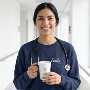 Puede incluir: Una sudadera azul marino con el nombre "Emily RN" y un diseño de estetoscopio. Se sostiene una taza blanca con un diseño de media luna. La persona sonríe y lleva un estetoscopio alrededor del cuello.