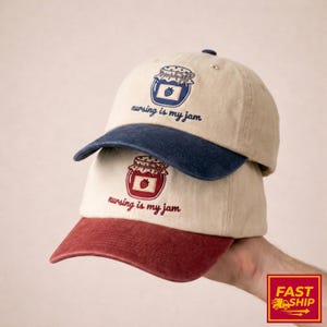 Puede incluir: Dos gorras de béisbol beige apiladas. La gorra superior tiene una visera azul marino, y la inferior una visera granate. Cada gorra presenta un diseño de tarro bordado con el texto "nursing is my jam".