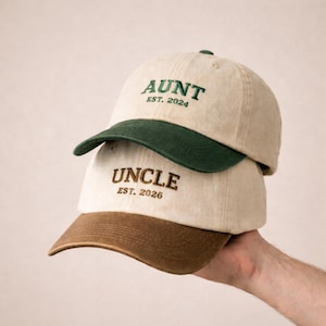 Op de afbeelding: Twee baseballpetten op elkaar gestapeld. De bovenste pet is beige met een groene klep en het woord "AUNT" geborduurd in groen, met "EST. 2024" eronder. De onderste pet is beige met een bruine klep en het woord "UNCLE" geborduurd in bruin, met "EST. 2026" eronder.